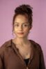 Portrait of a young woman with curly hair against a pink background, wearing a brown shirt.