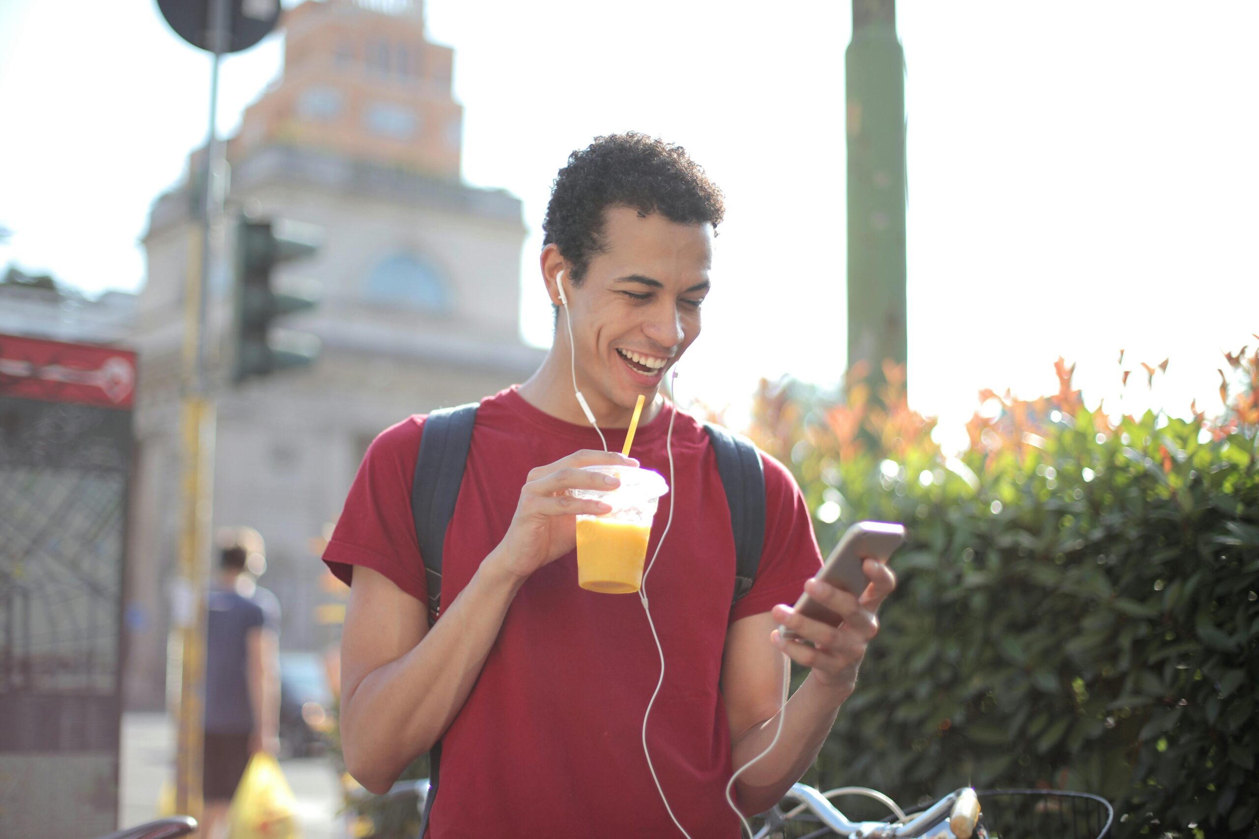 Services Happy ethnic guy in casual wear and with backpack having cup of fresh juice using smartphone while standing on street and listening to music