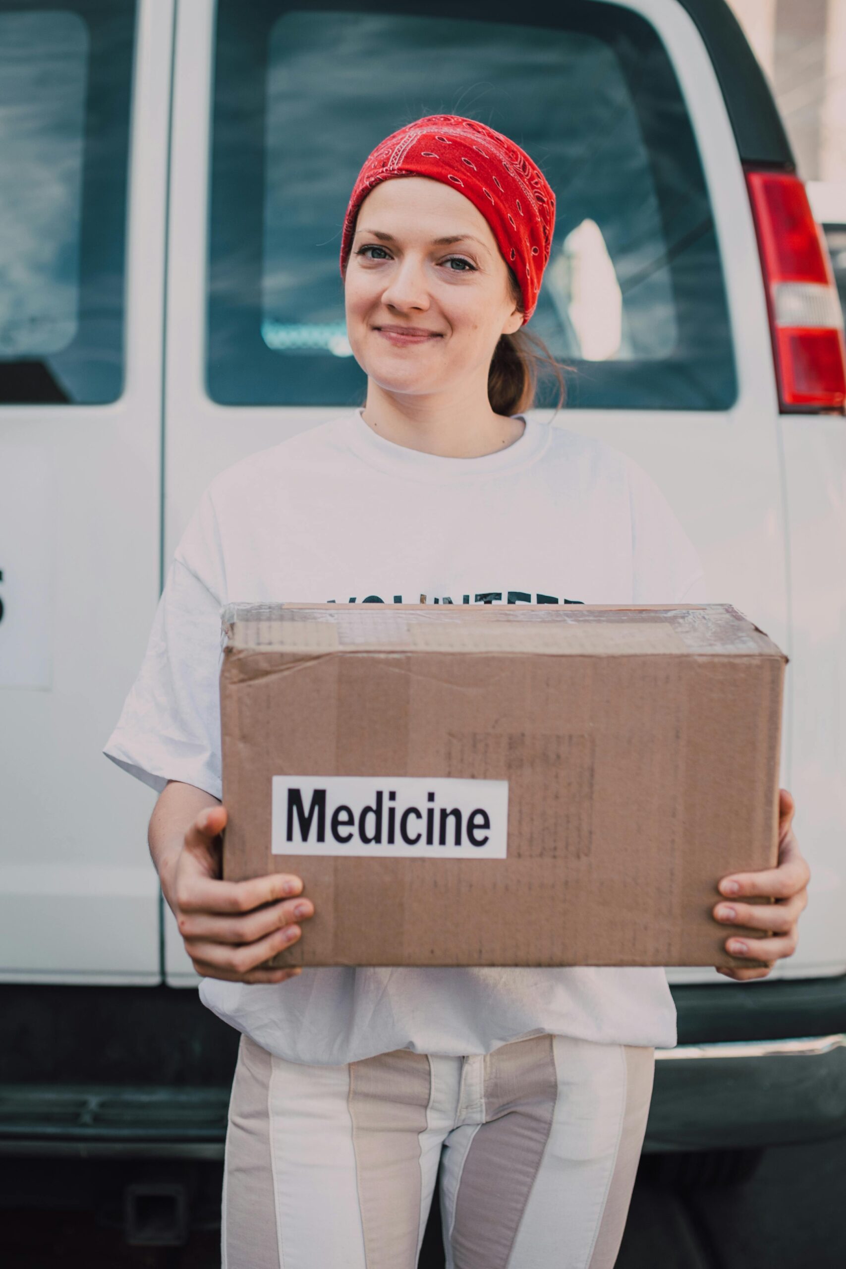 About A smiling female volunteer holding a donation box labeled 'Medicine' in front of a van.