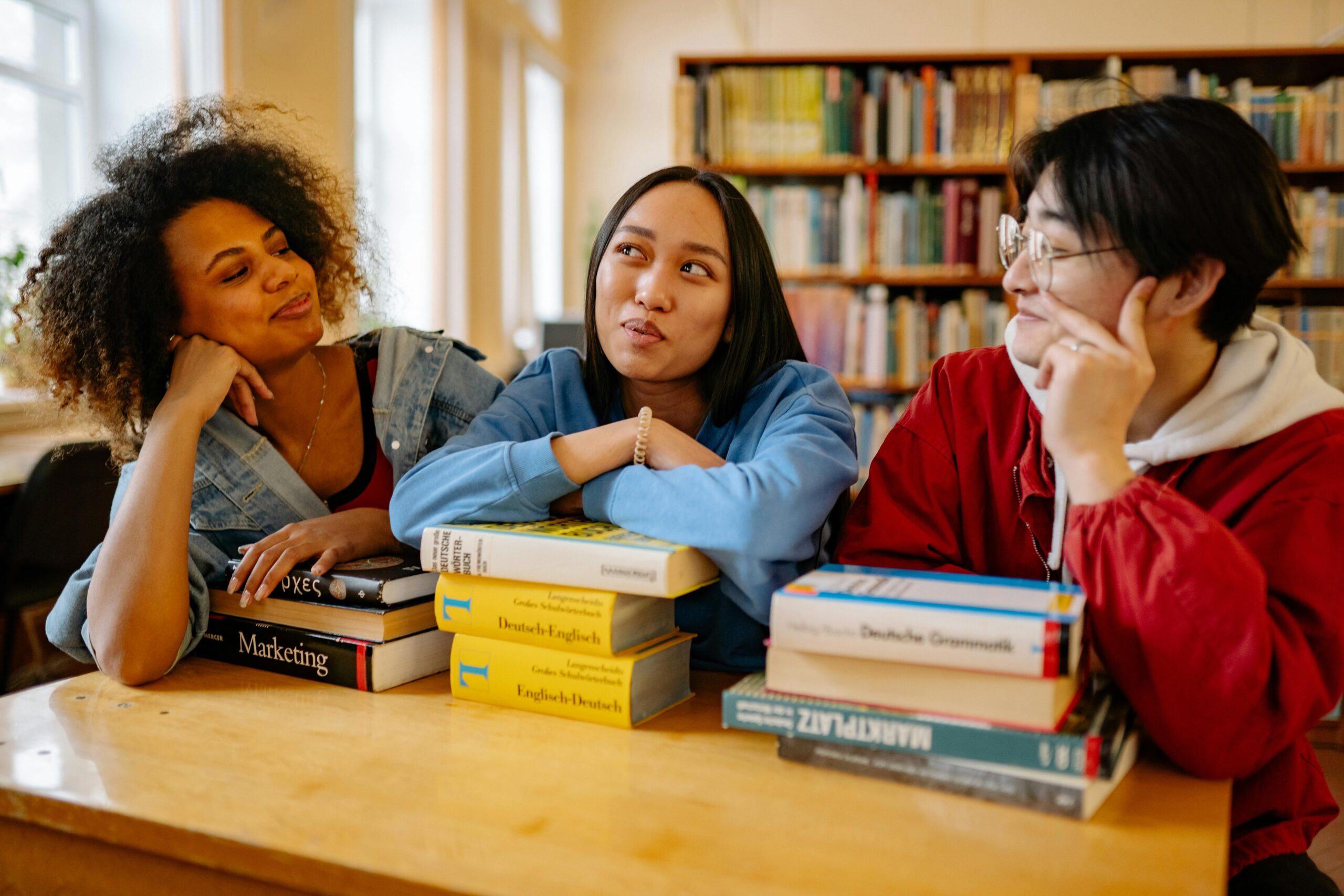 Services Three diverse students studying together in a university library with books around them.